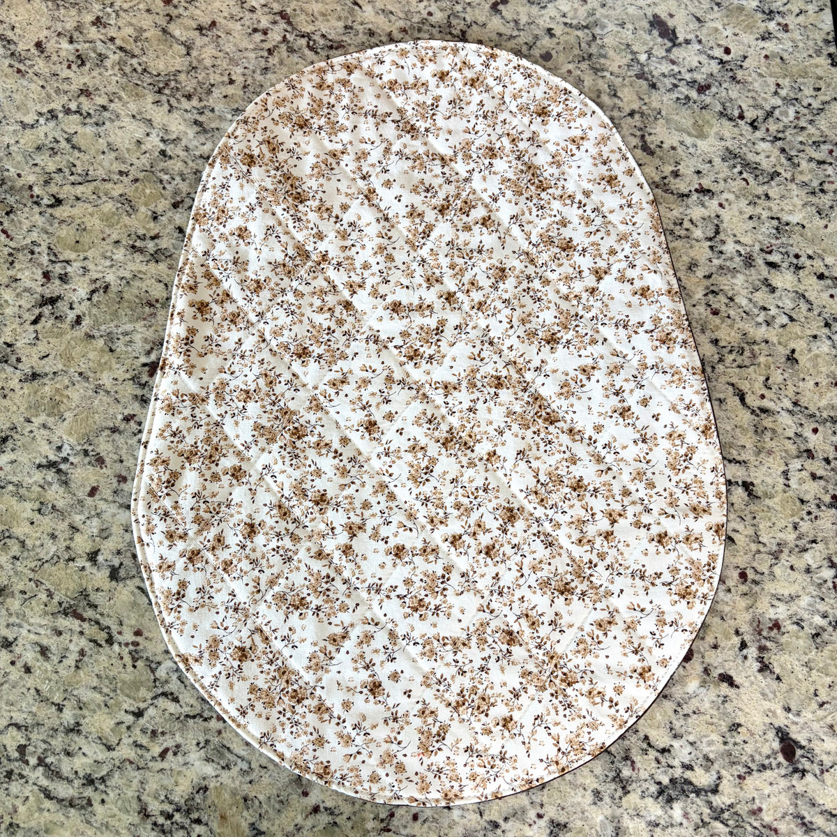 An oval-shaped fabric mat, featuring a white background with a dense brown floral pattern, laid flat on a speckled granite countertop.
