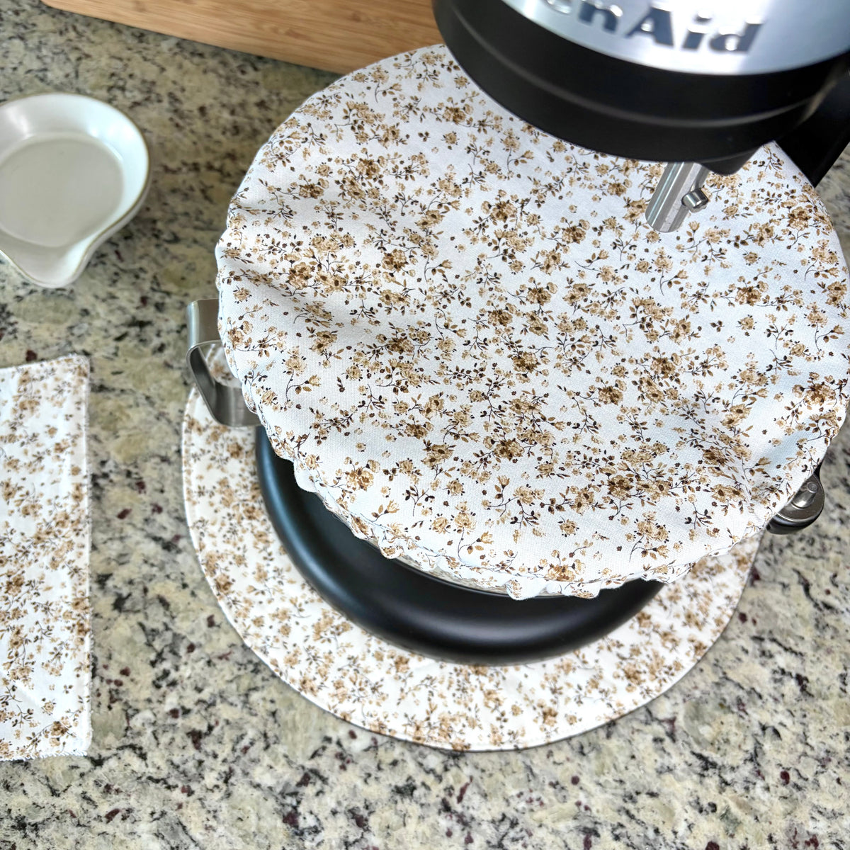 A close-up of a stand mixer with a brown floral  bowl cover, set on a matching mat on a granite countertop with a small white spoon rest nearby.