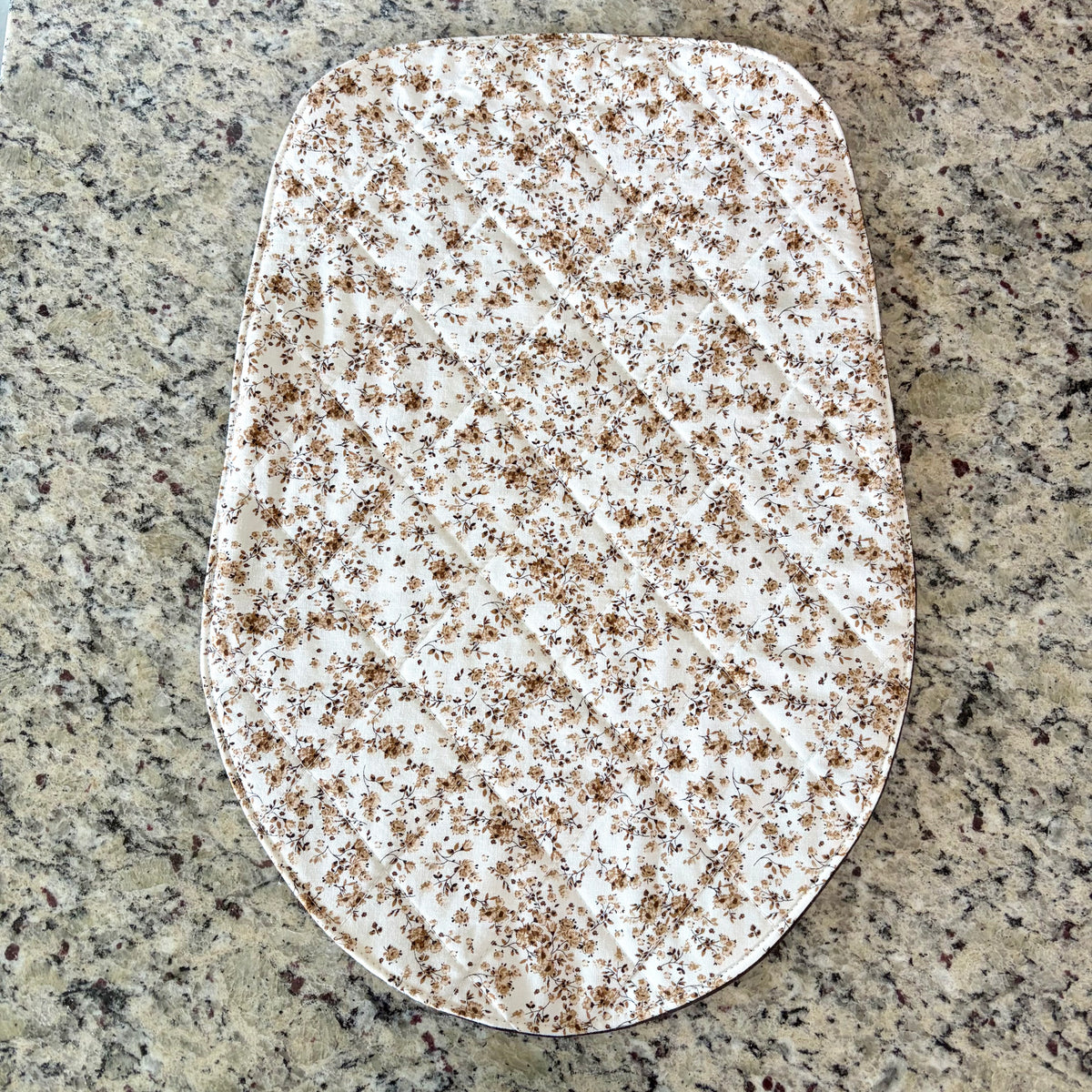 A dense floral white and brown fabric  mat with a rounded bottom, laid flat on a speckled granite countertop.