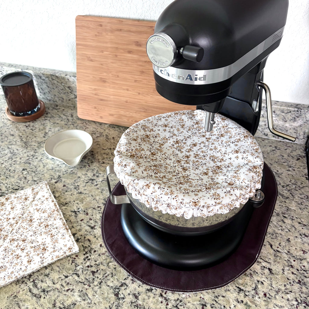 A black KitchenAid stand mixer with a white and brown floral fabric cover on a dark brown mat, set on a granite countertop next to a matching cloth and a wooden cutting board.