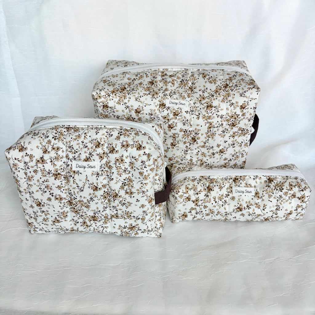 Three floral zippered pouches in different sizes arranged on a white fabric backdrop. The pouches have a cream background with a dense brown and beige floral print, and a small white fabric brand label on the front.