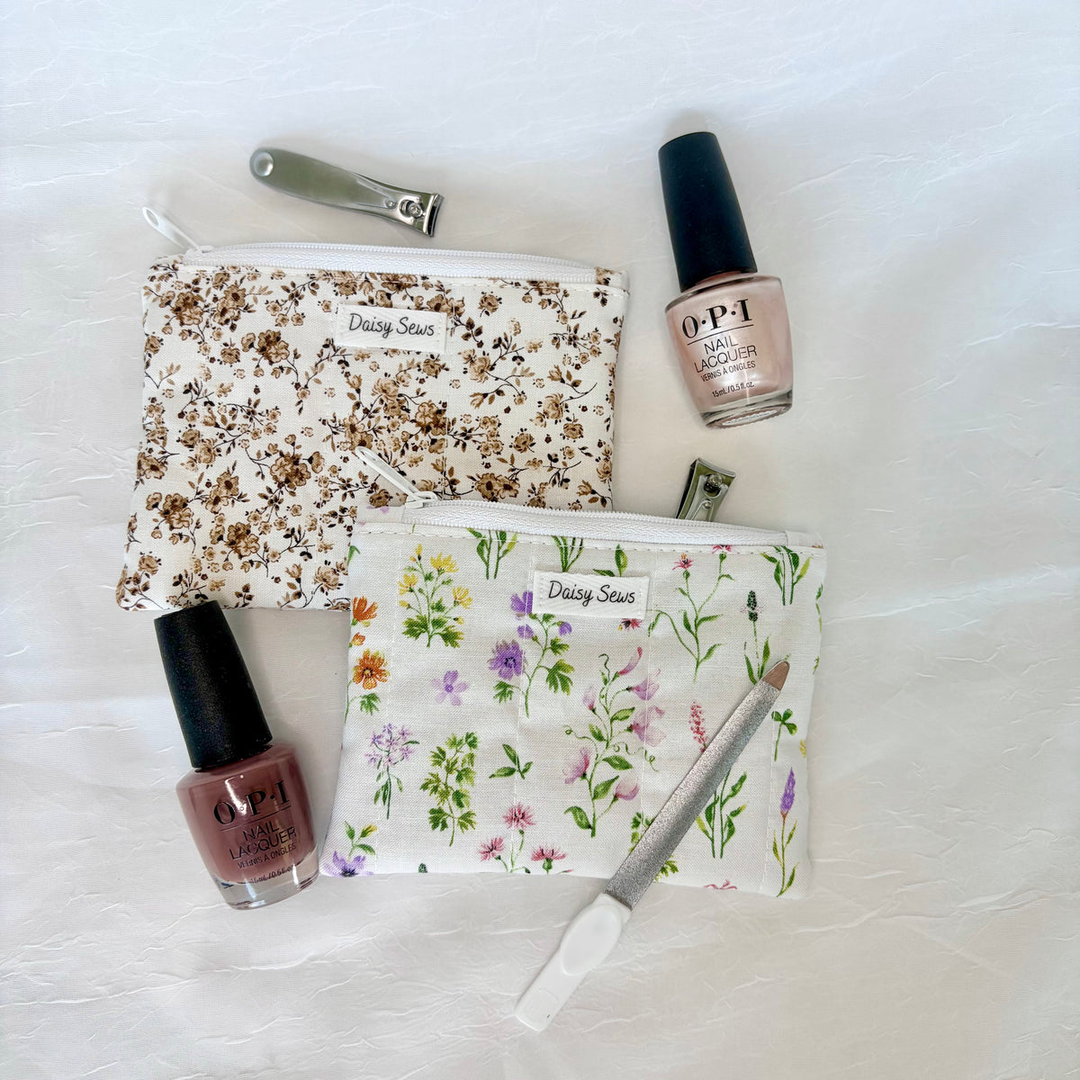 Two floral zippered cosmetic pouches with nail care accessories arranged on a white textured fabric background. One pouch has a brown floral print and the other has a multicolor wildflower print.