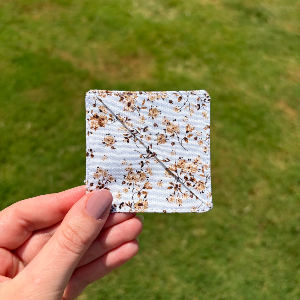 A hand holding a square fabric patch with a white background and a beige and brown floral pattern, set against a blurred green grass background.