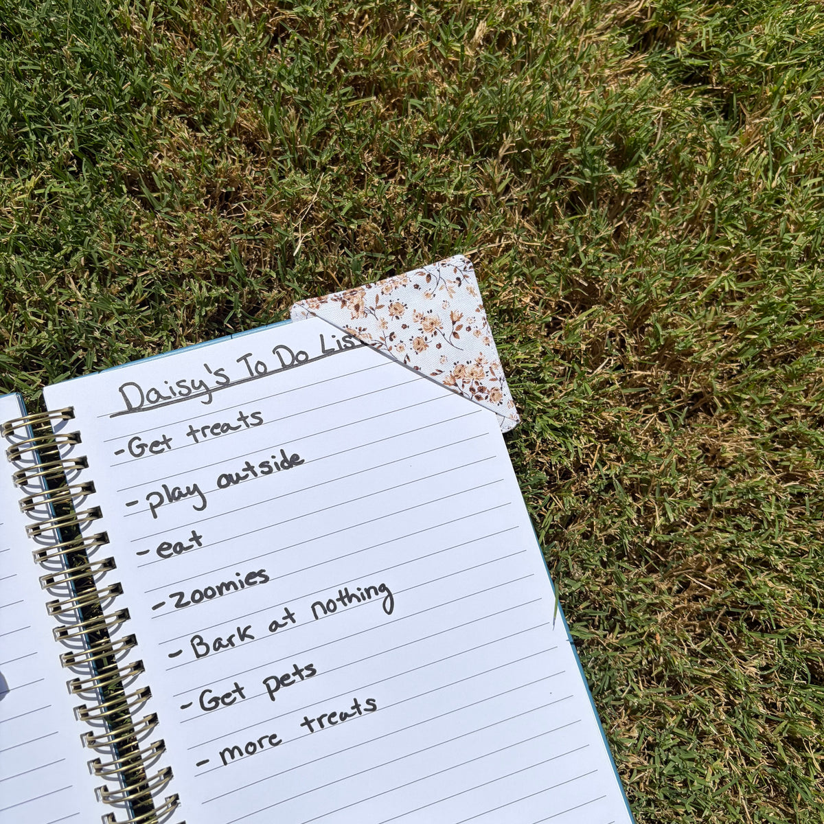 A spiral-bound lined notebook lies open on green grass, with handwritten text reading 'Daisy's To Do List' and a small decorative bookmark or page tab attached. No person or animal is visible, but the real outdoor grass environment makes this Lifestyle rather than Product/Packaging.