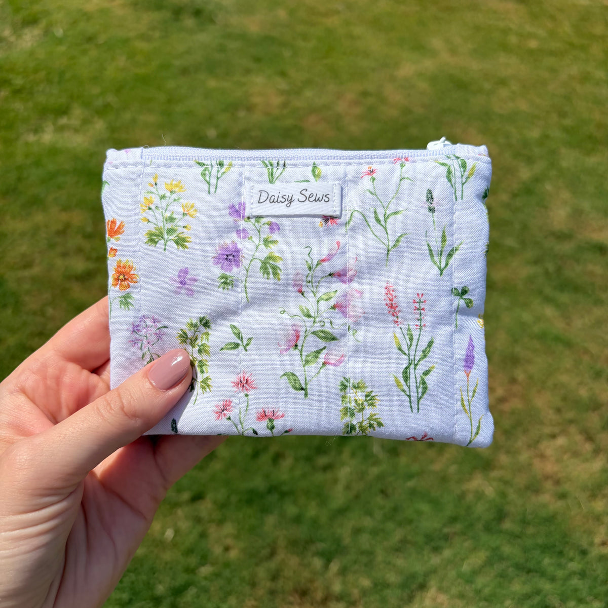 A hand holds a small zippered pouch with a light blue floral print. The pouch has a white fabric label that reads 'Daisy Sews' and is photographed outdoors against a blurred green grass background.