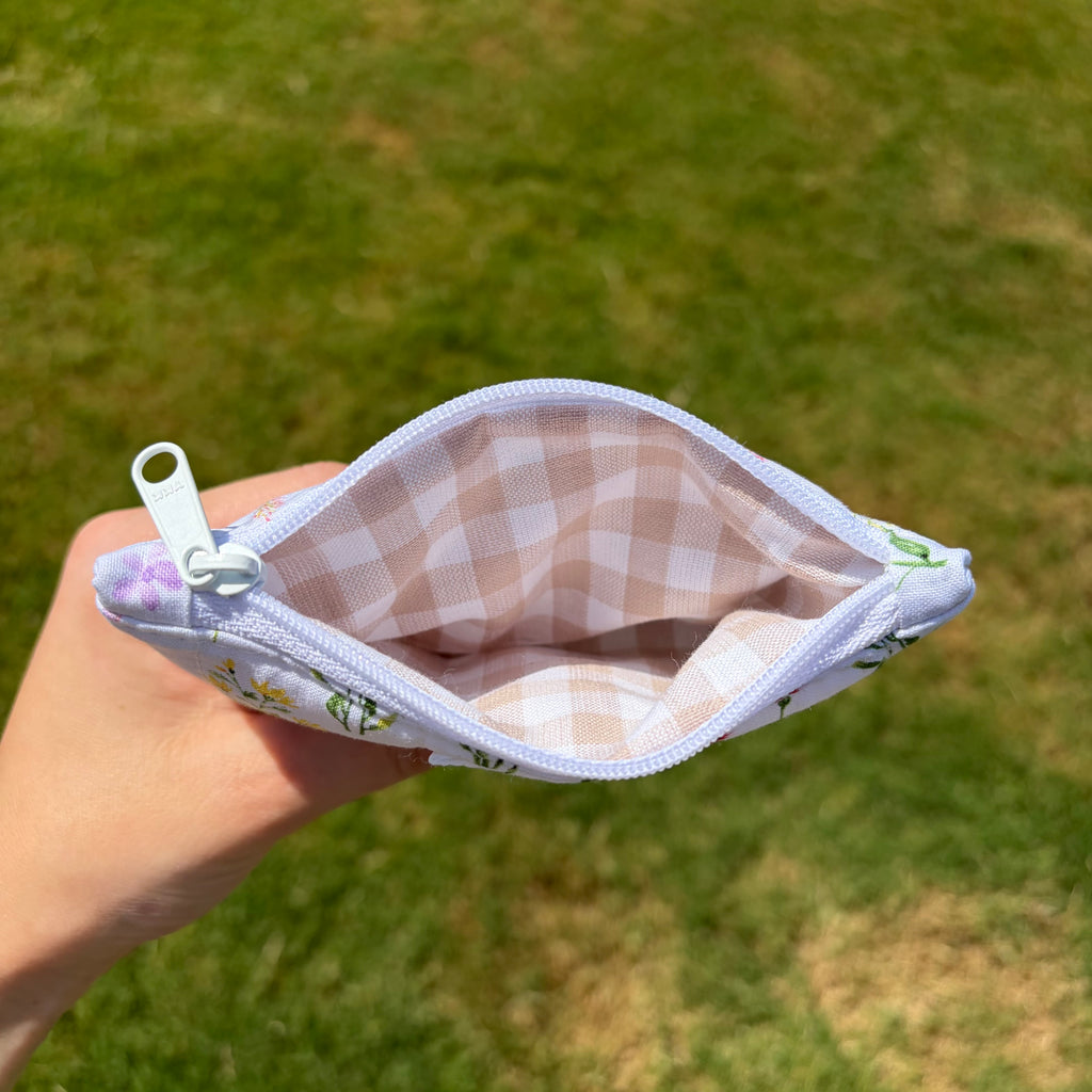 A hand holding an open zippered pouch with a white and beige gingham patterned lining and a white floral exterior, set against a blurred green grass background.