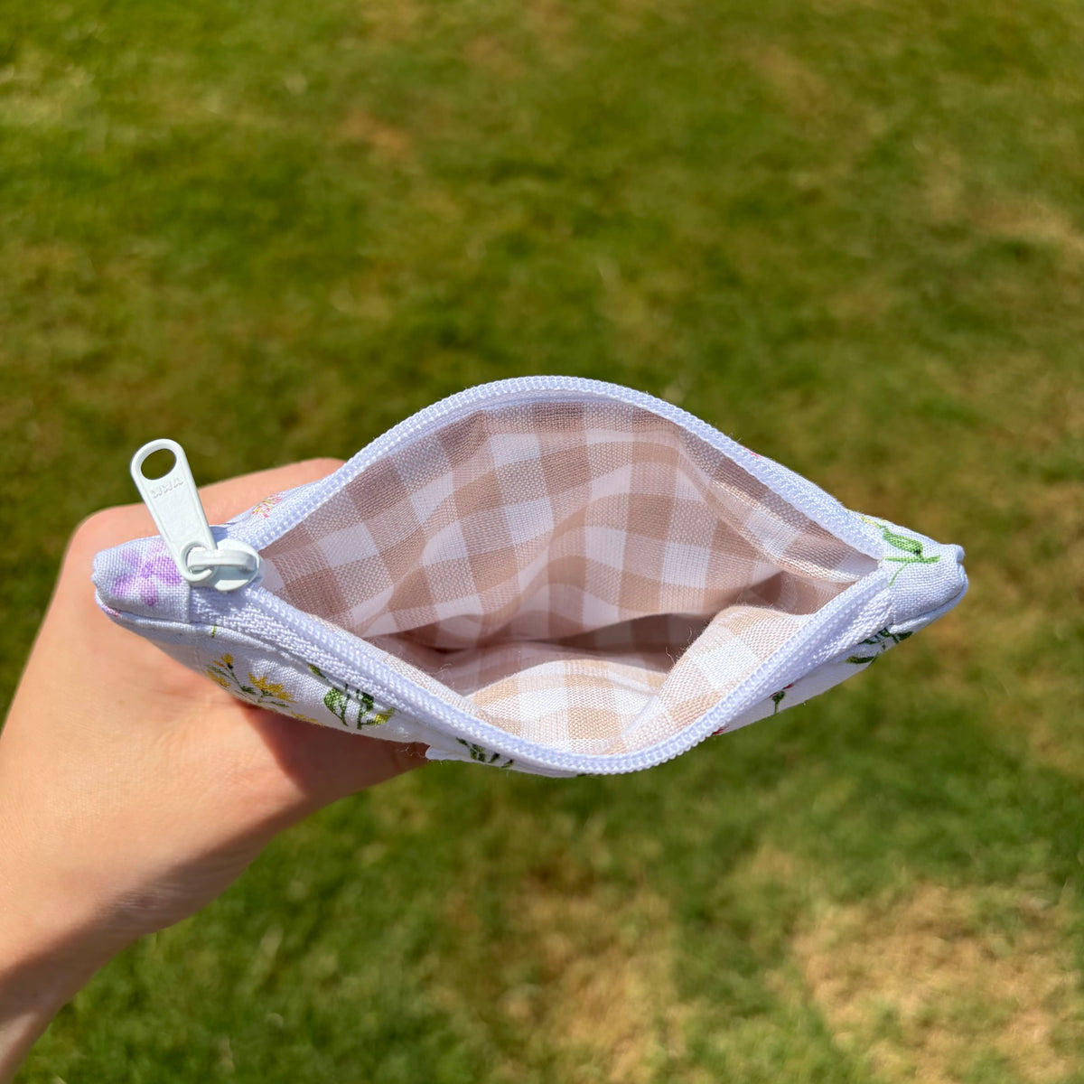 A hand holding an open zippered pouch with a white and beige gingham patterned lining and a white floral exterior, set against a blurred green grass background.