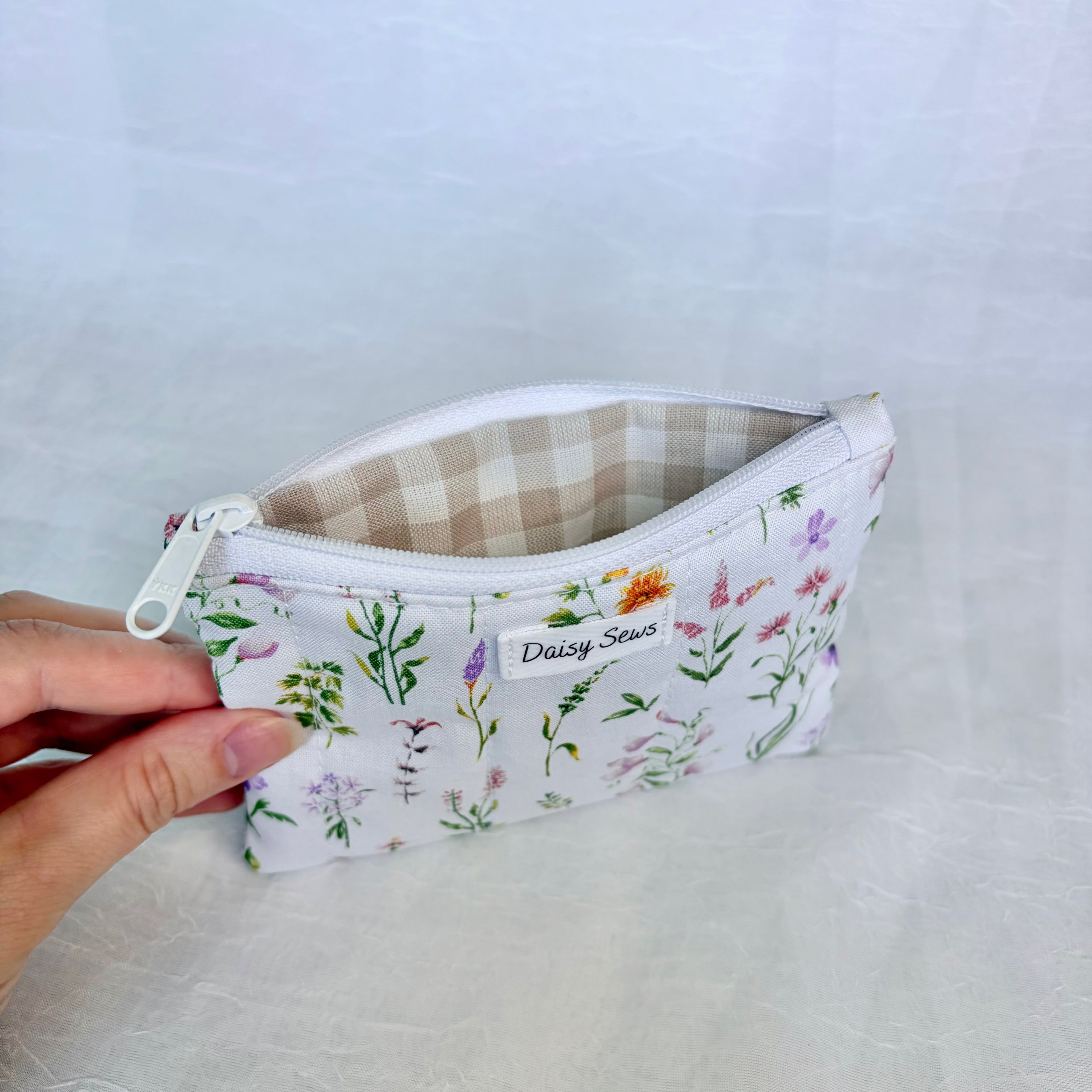 Floral-patterned pouch with beige gingham held by a hand on a light gray background