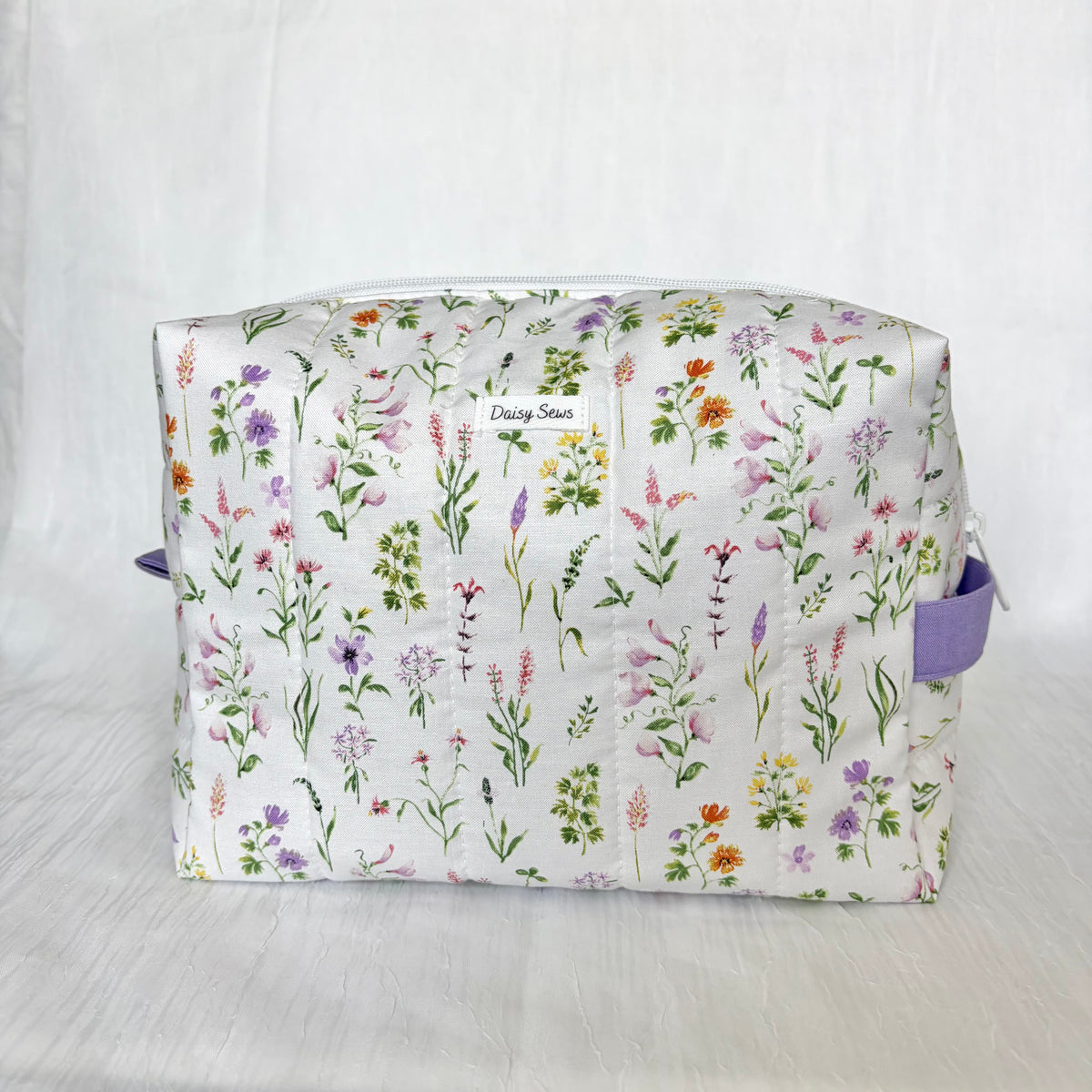 A quilted cosmetic pouch with a white background and a colorful watercolor wildflower print, featuring purple side handles and a small 'Daisy Sews' label.