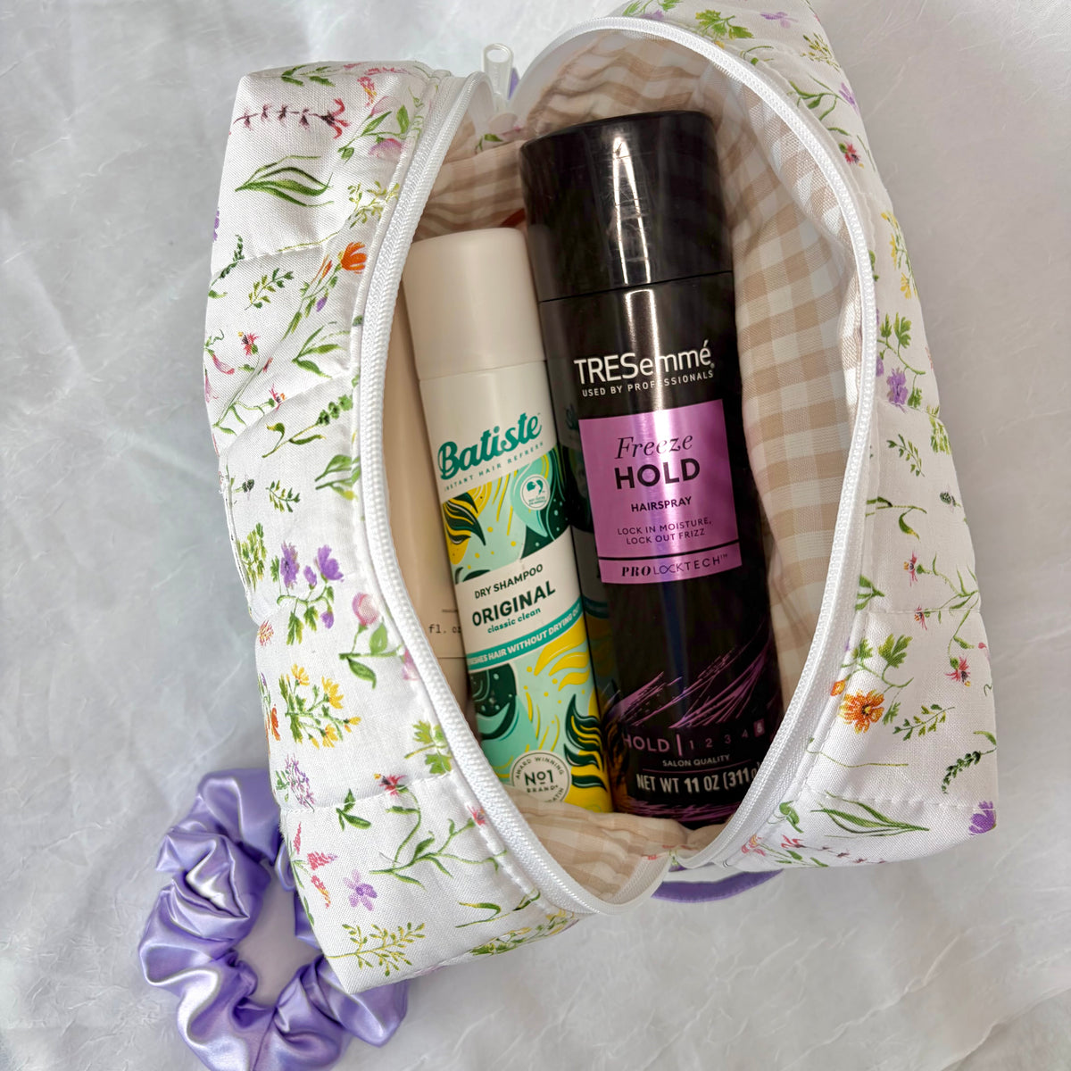 A floral zippered cosmetic bag filled with toiletries, including a bottle of Tresemme Freeze Hold hairspray and a dry shampoo, on a white textured fabric background with a purple scrunchie nearby.