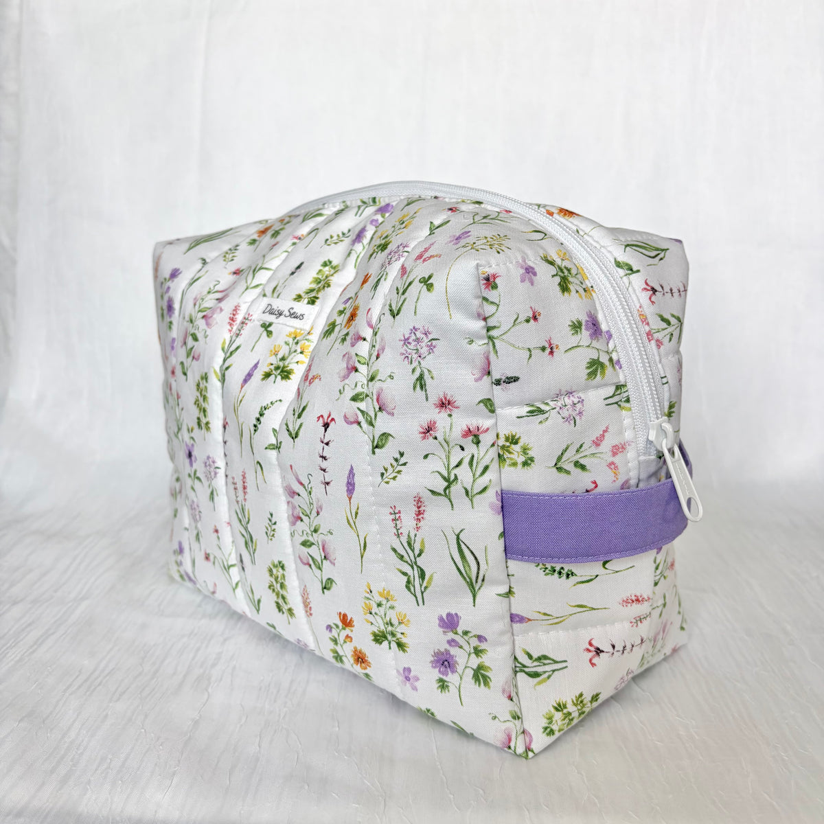A rectangular zippered cosmetic pouch with a white background and a colorful wildflower print, featuring a purple fabric loop handle on the side.