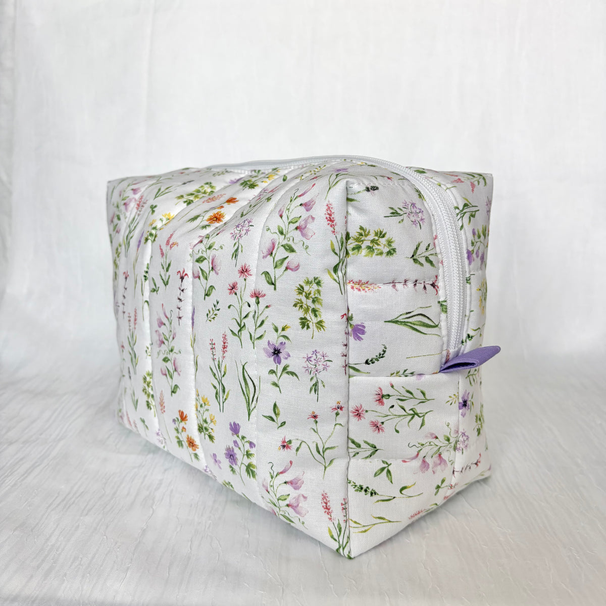 A rectangular zippered pouch with a white background and a delicate multicolor floral print, featuring a small purple zipper pull, set against a white fabric backdrop.