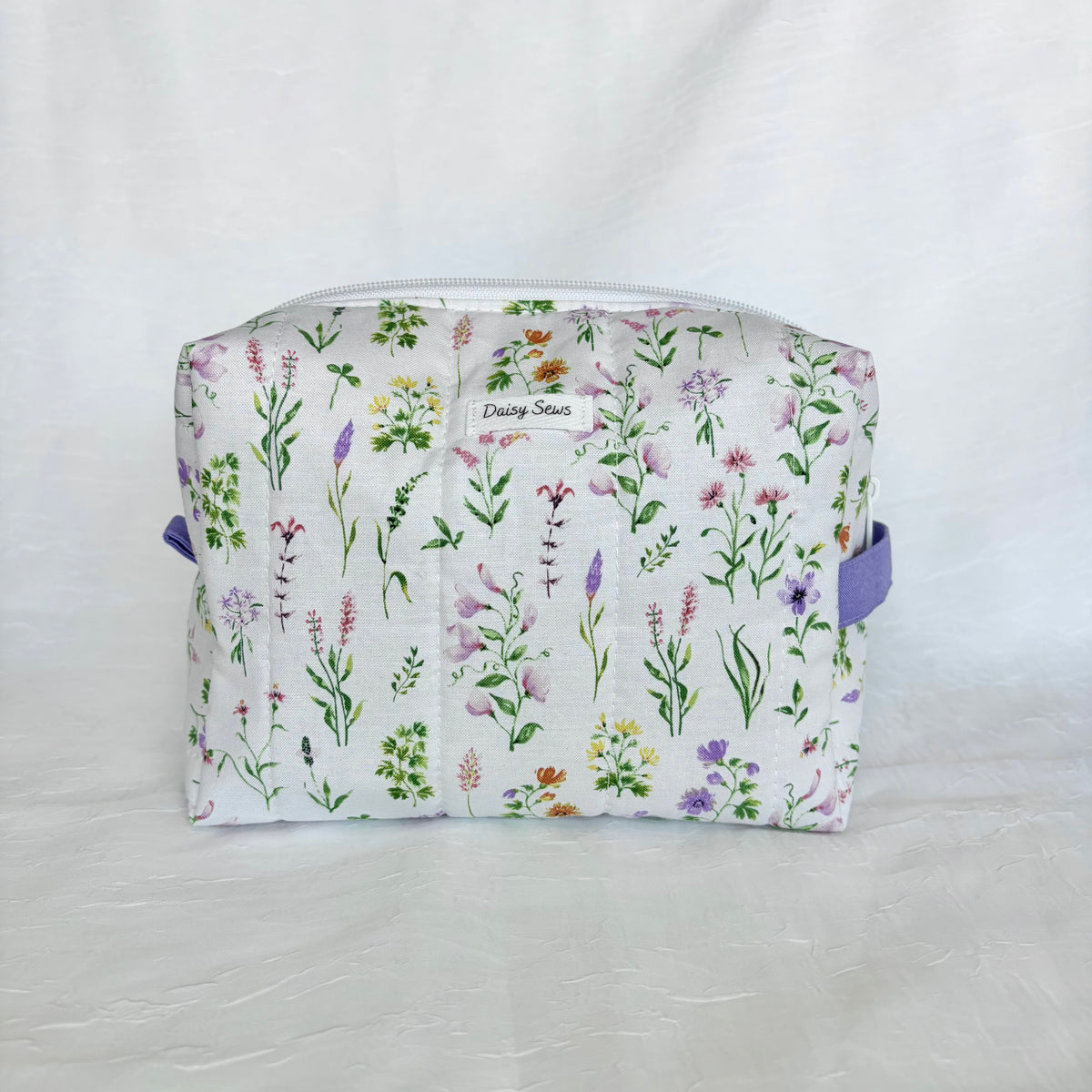 A quilted fabric cosmetic pouch with a white background and a colorful wildflower print, featuring purple side handles and a small  'Daisy Sews' label.