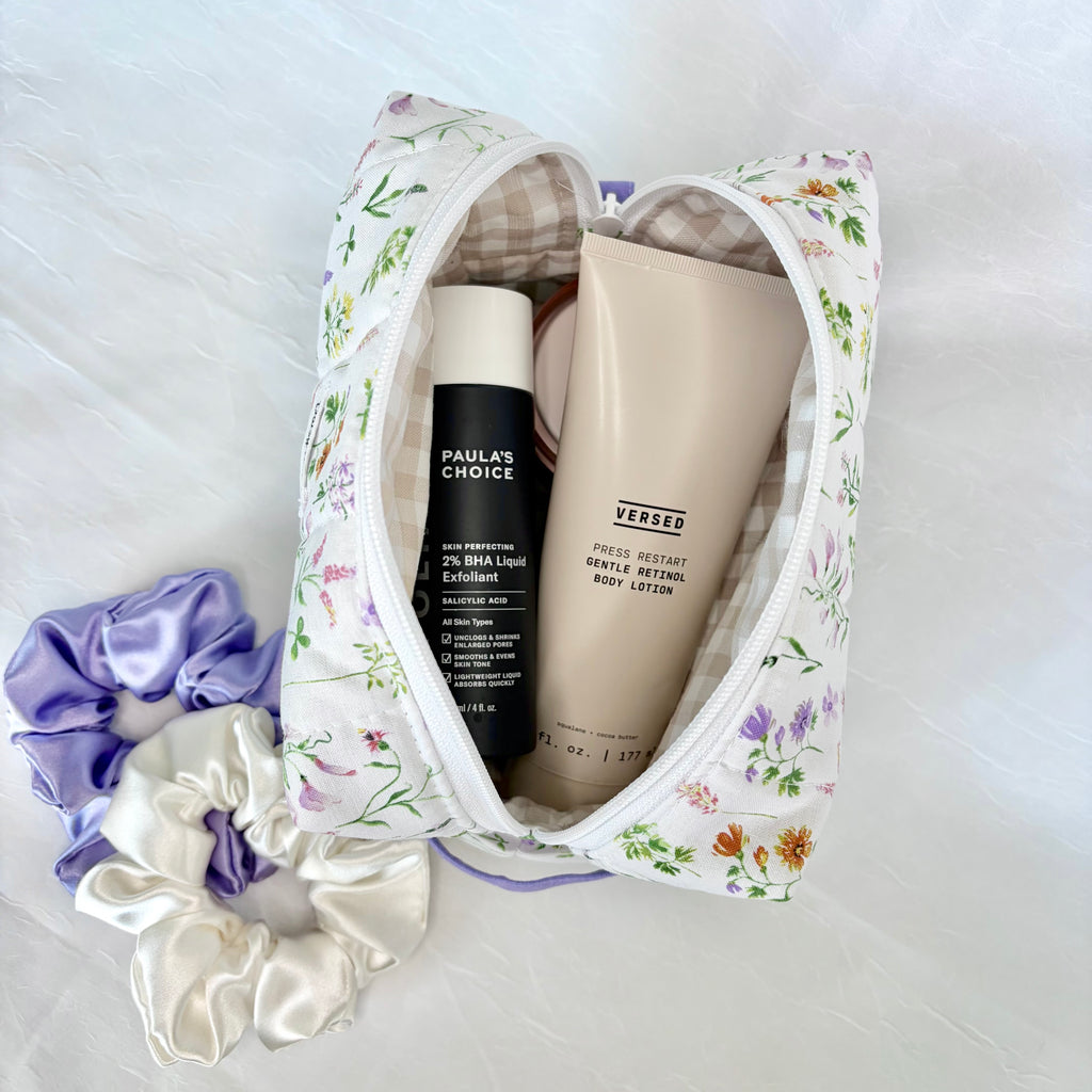 A floral zippered travel or vanity bag filled with toiletries, including a black bottle labeled Paula's Choice and a beige tube labeled Versed, arranged on a light fabric surface with purple and white satin scrunchies beside it.