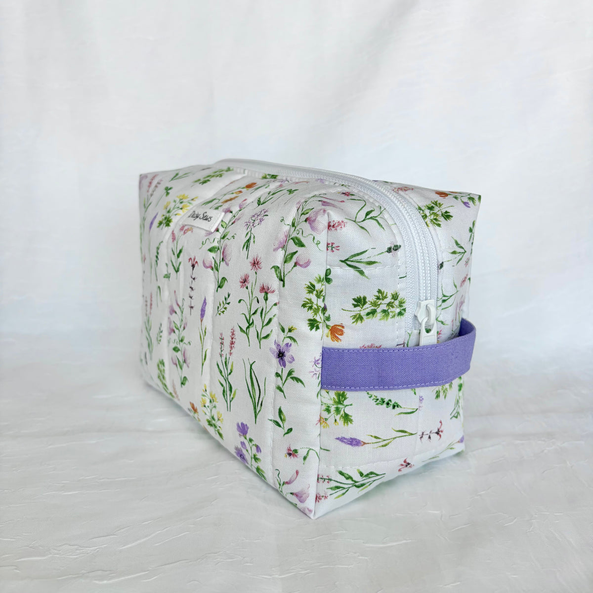 A rectangular zippered cosmetic pouch with a white background and a colorful wildflower print, featuring a purple fabric handle, set against a white fabric backdrop.