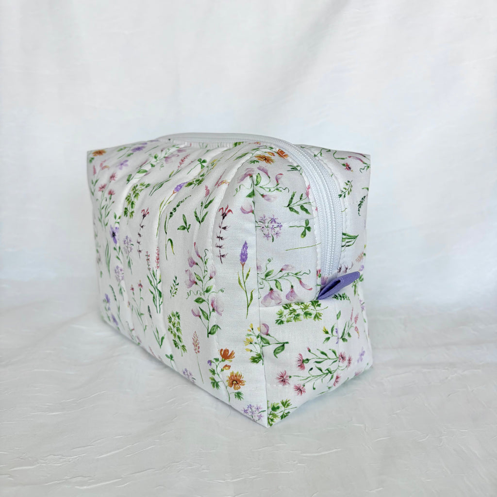 A rectangular zippered pouch with a white background and a delicate multicolor wildflower print, featuring a small purple zipper pull, photographed against a white fabric backdrop.