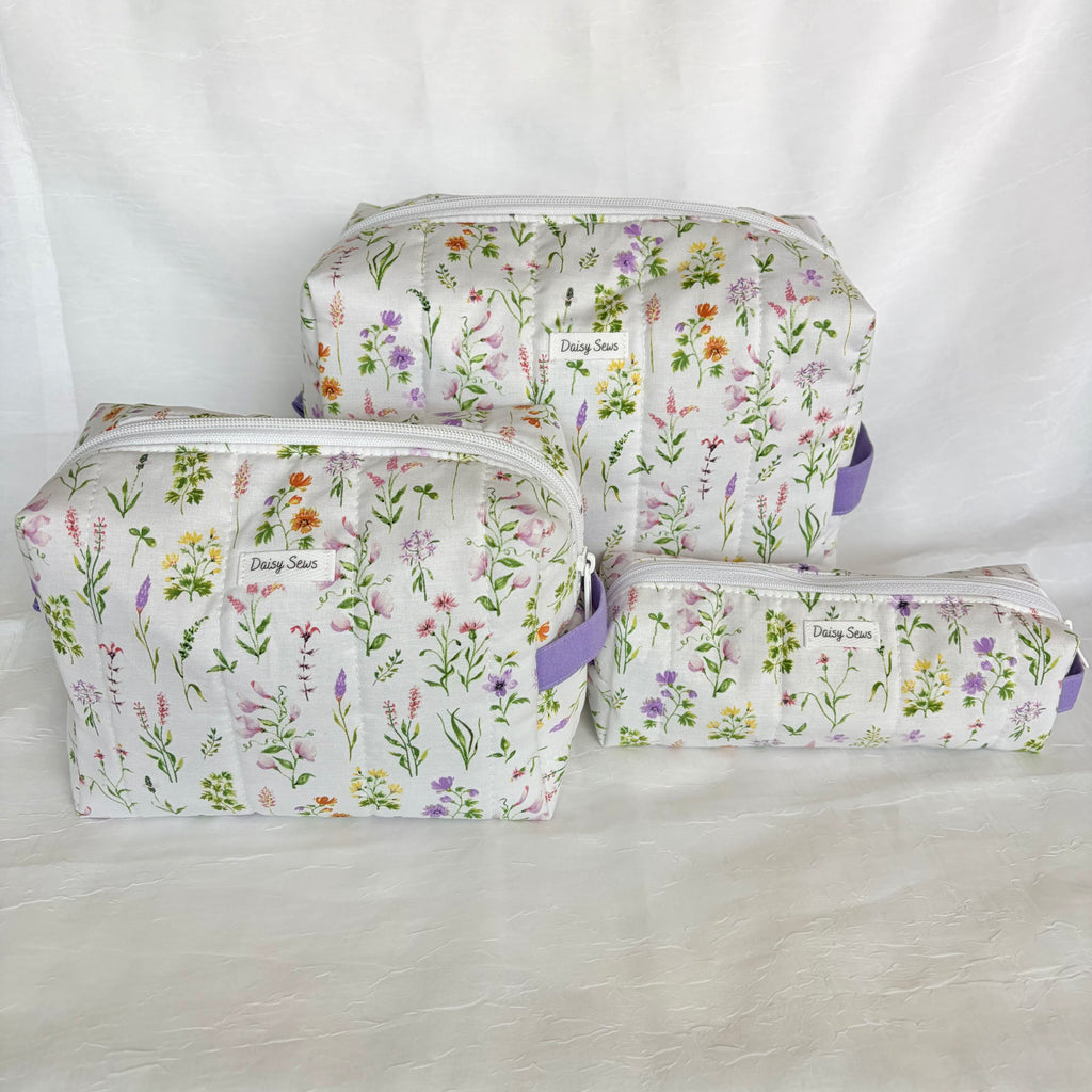 Three quilted cosmetic pouches in different sizes with a white background and a colorful wildflower print, featuring purple zipper pulls and a 'Daisy Sews' brand label.