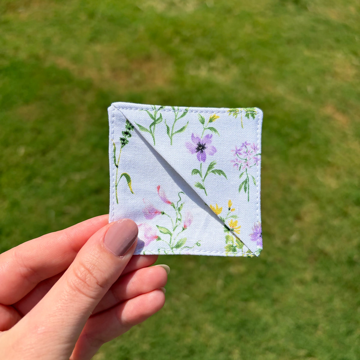 A hand with mauve-painted nails holds a small square fabric patch featuring a light blue background with a delicate purple, pink, and green floral print, set against a blurred green grass background.