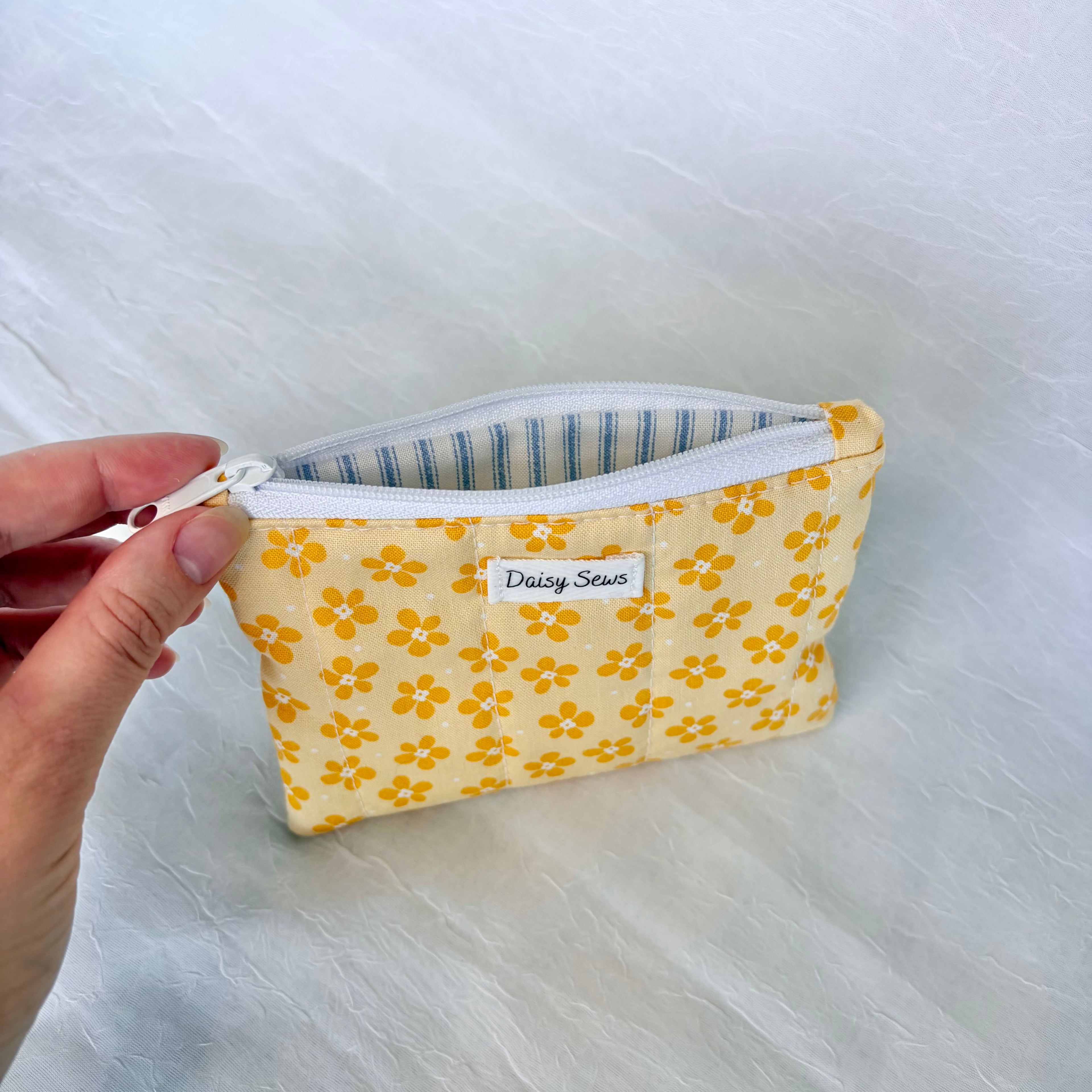 Yellow floral fabric pouch with a white zipper and blue striped liner held by a hand on a white background