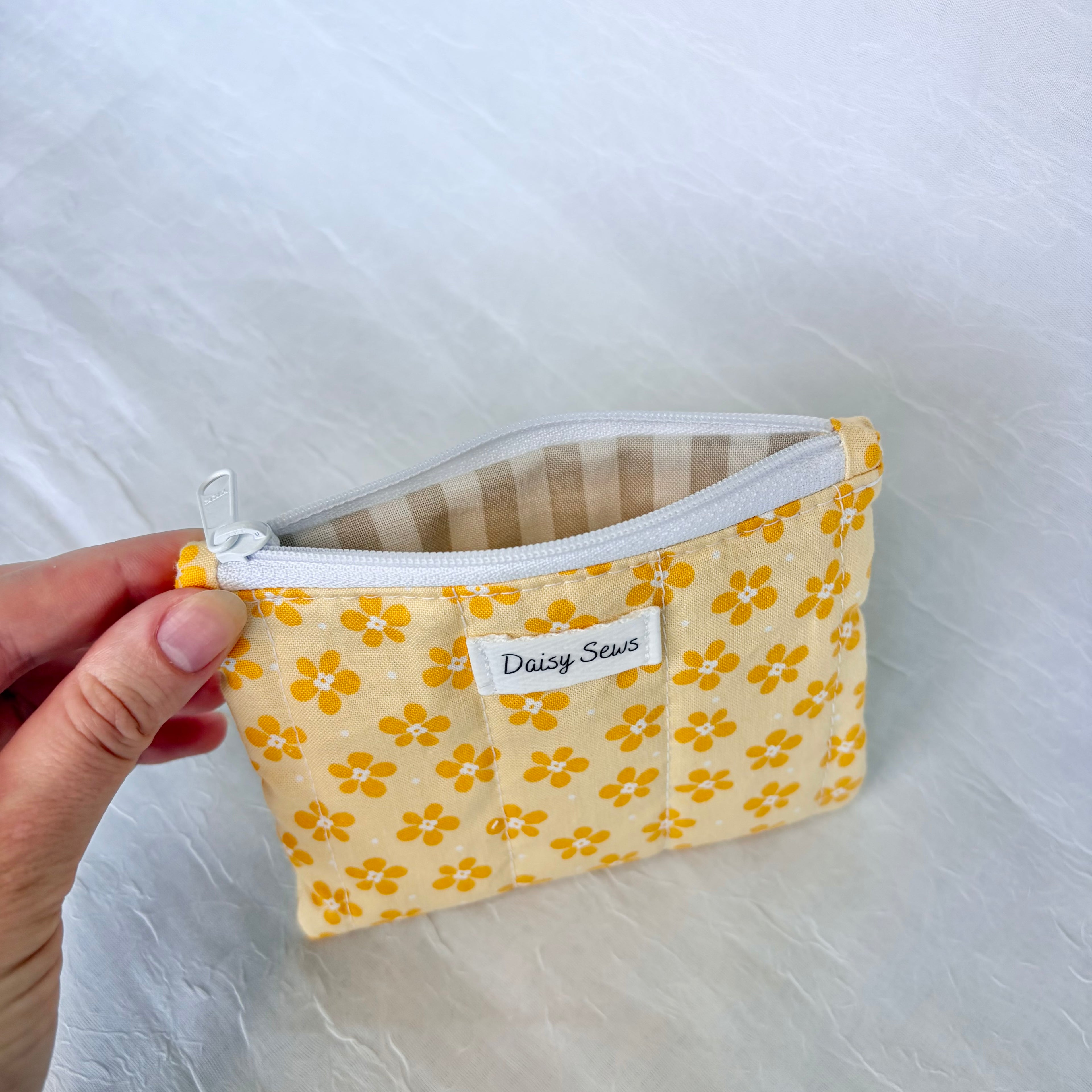 Hand holding a small fabric pouch with yellow floral pattern and tan striped liner and white zipper, on a light background.
