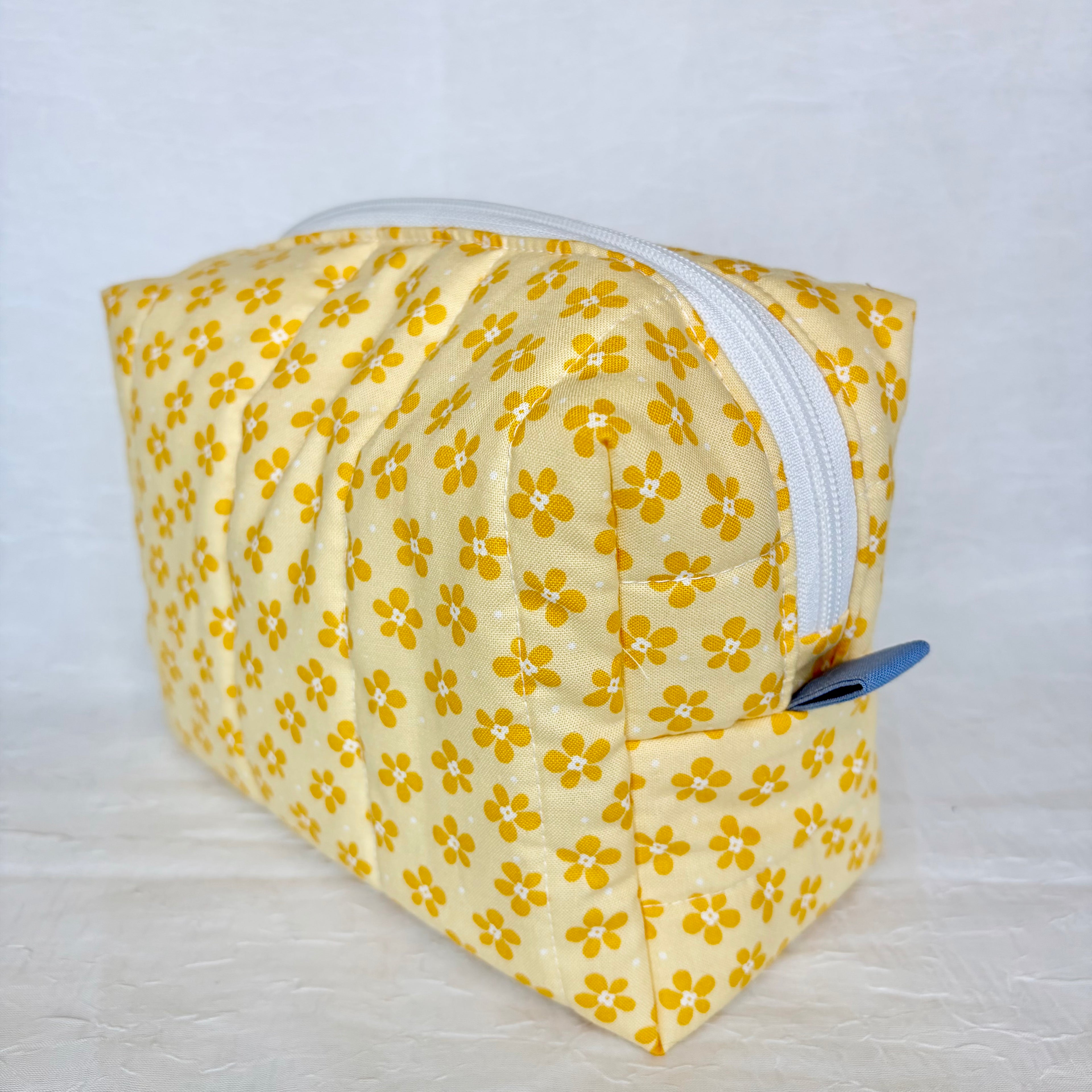 Yellow floral-patterned fabric bag with a white zipper and blue tab on a light gray background