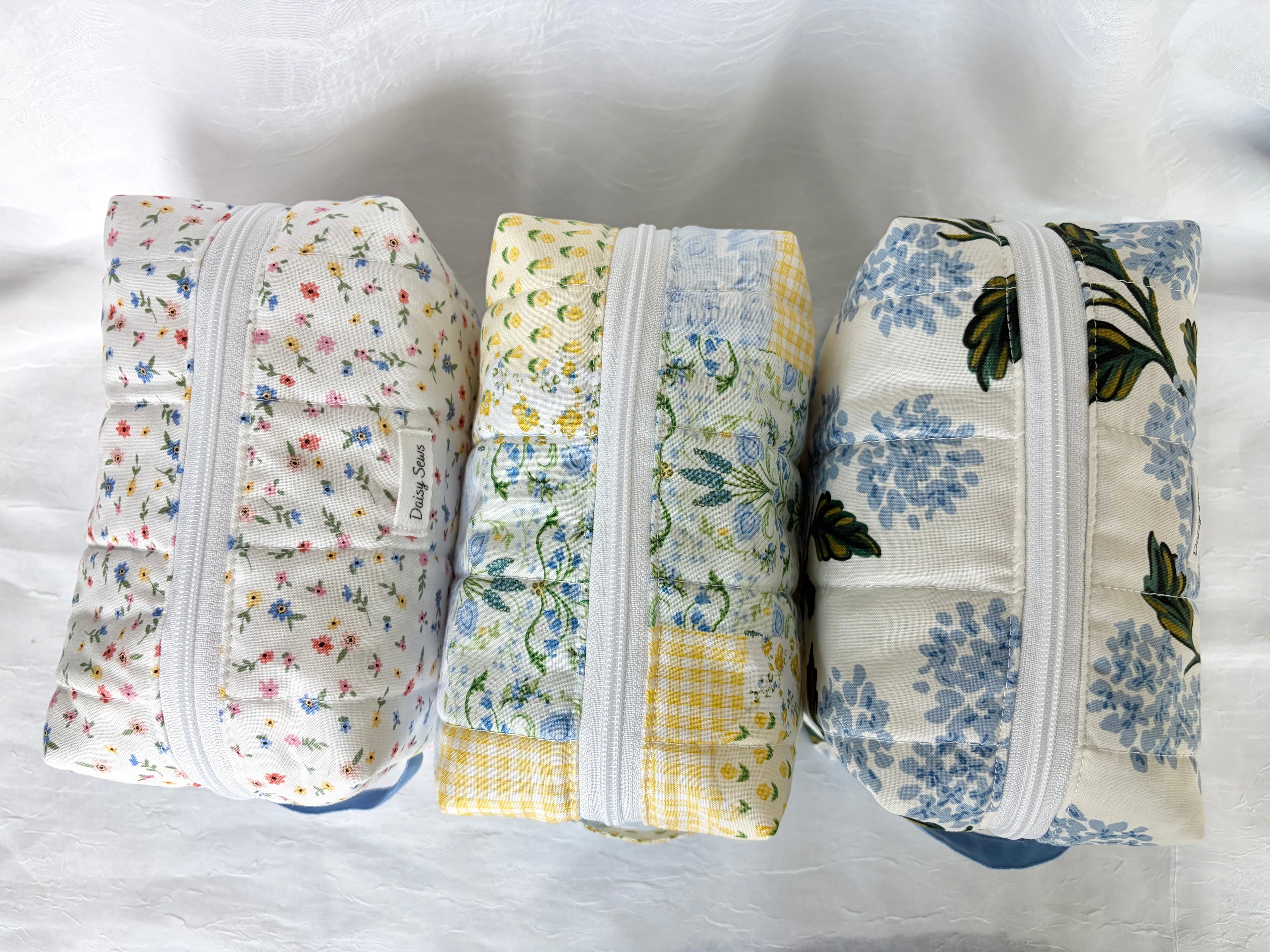 Top View of Three floral-patterned toiletry bags in blue, yellow, pink and white colors with white zippers on a white background