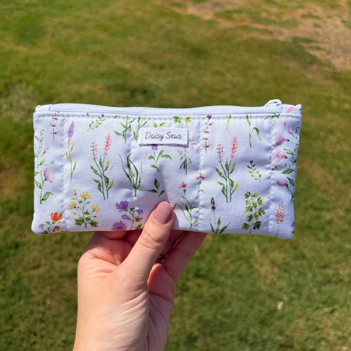 A hand holding a light blue zippered pouch with a watercolor-style wildflower print, set against a blurred green grass background. The pouch features a white fabric label with the brand name 'Daisy Sews'.