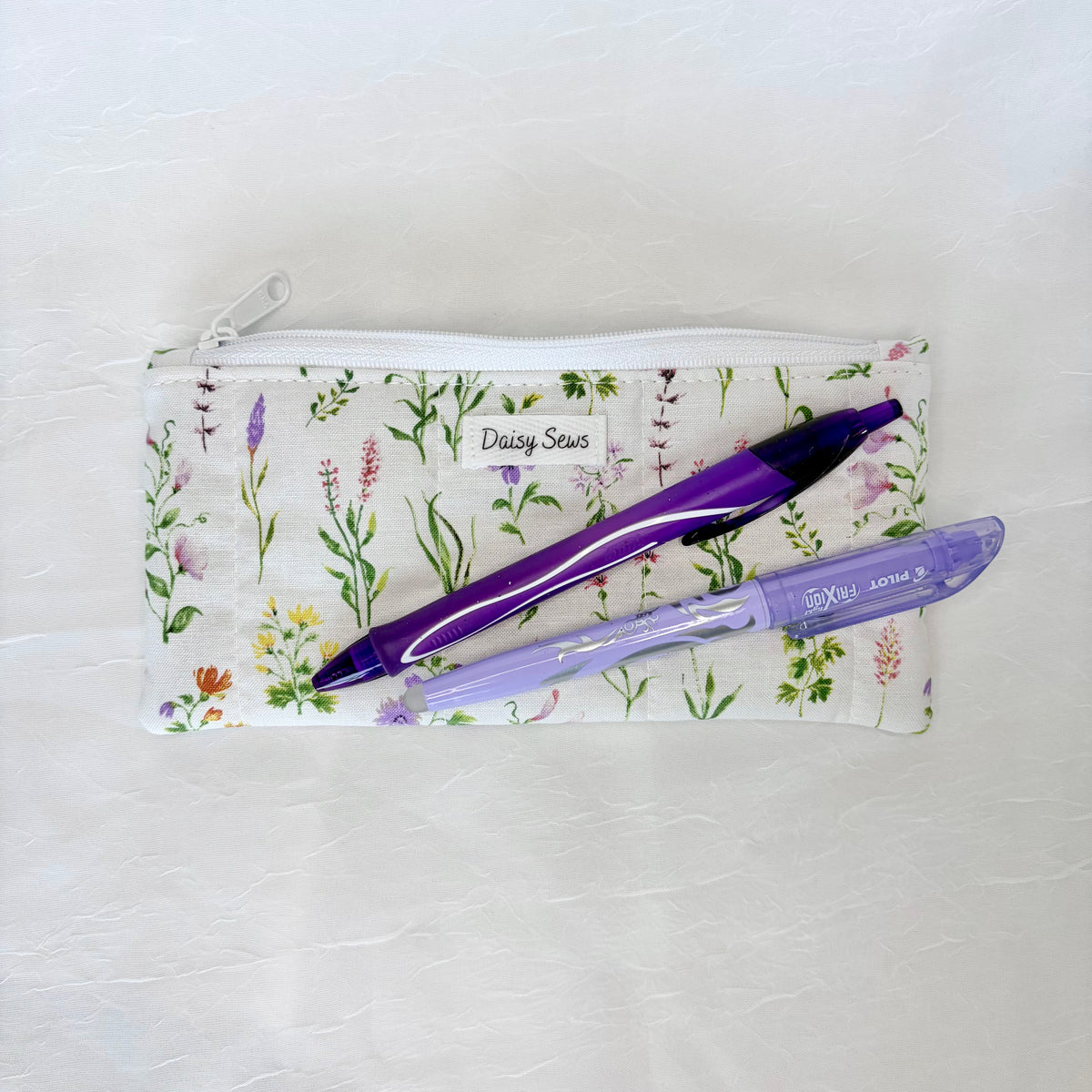 A small zippered pouch with a white background and a delicate multicolor wildflower print, featuring a white fabric label that reads 'Daisy Seeds'. Two purple pens are placed on top of the pouch.