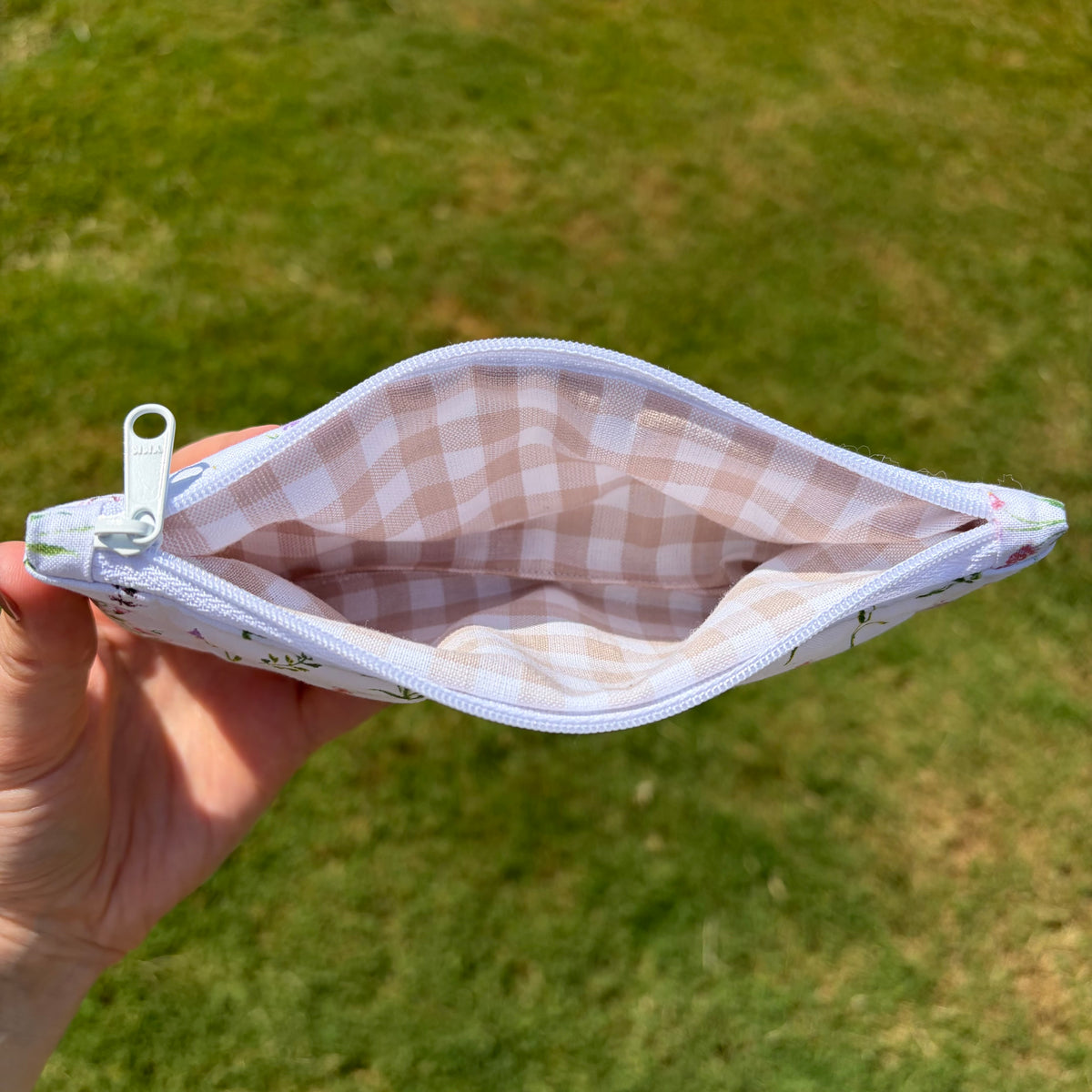 A hand holding open a small zip pouch outdoors over a grassy lawn. The pouch has a white exterior with a subtle floral print and a light pink and white gingham lining.